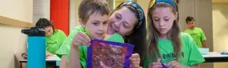 A U N E student compliments two children's artworks made during Autism Camp